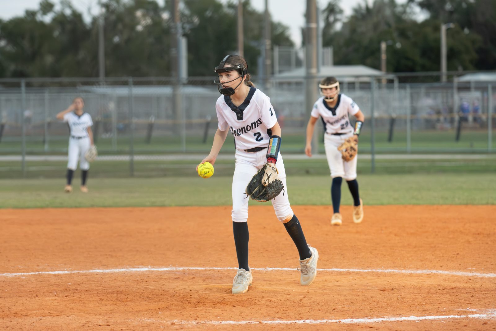 Camden delivering a pitch in a white Phenoms uniform with fielders behind her